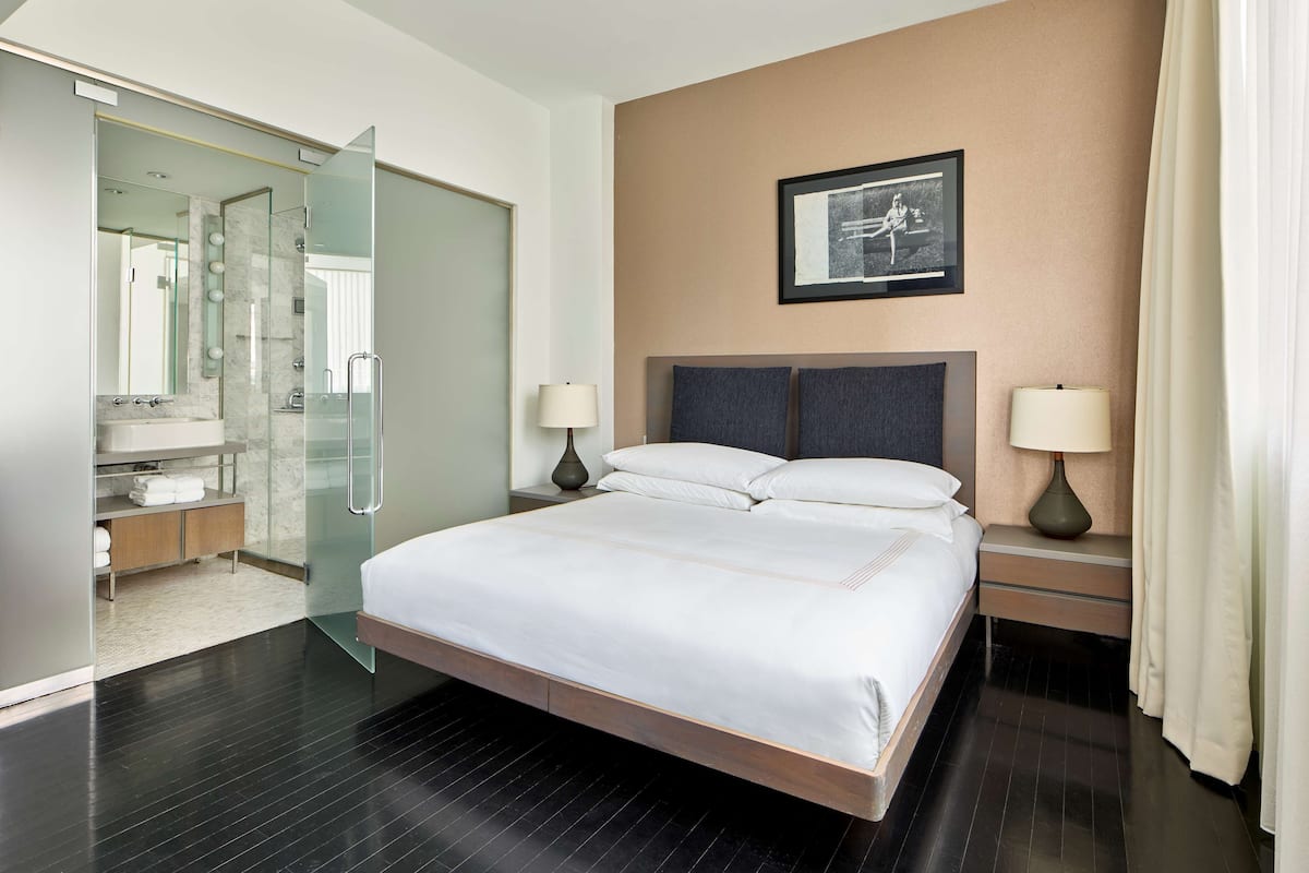 A modern hotel room with a large bed, bedside lamps, a framed picture on the wall, and a glass shower enclosure.