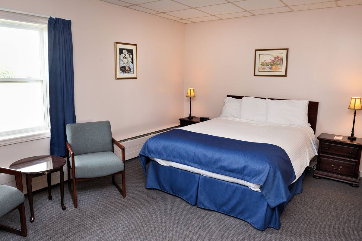 Un cuarto de hotel con una cama, dos mesitas de noche con lámparas, una silla y dos cuadros en las paredes.