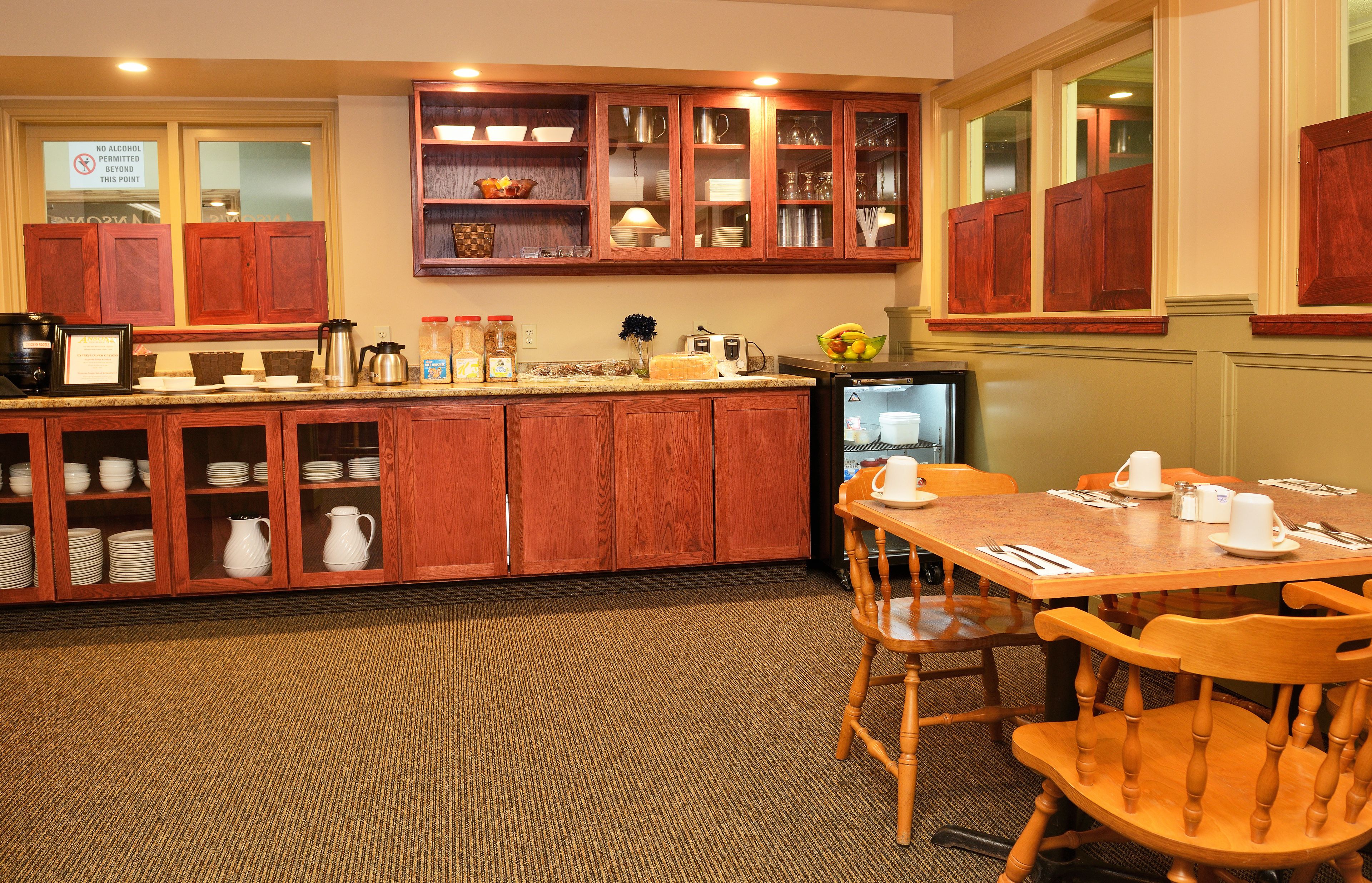 A hotel kitchen with wooden cabinets, a coffee station, and a dining area with chairs and a table set for two.
