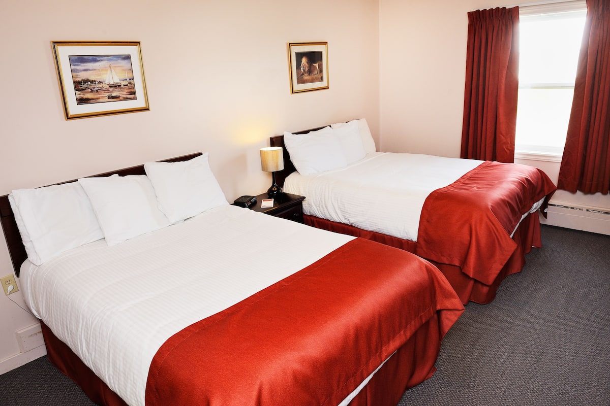 Habitación de hotel con dos camas, una mesita de noche, una lámpara y dos cuadros en la pared.