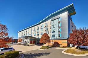 A modern hotel building with a covered entrance, multiple floors, and a parking lot with cars. - Aloft by Marriott Dulles Airport North (Ashburn)