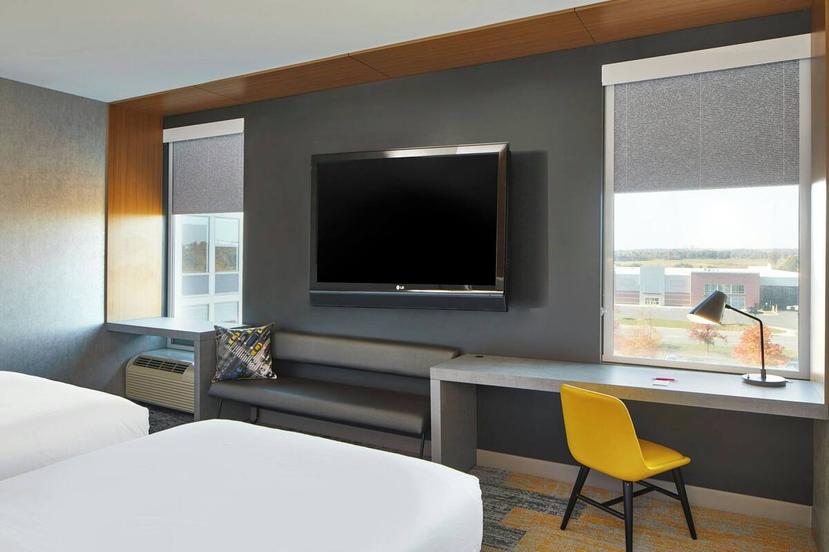 A hotel room with a large flat-screen TV mounted on the wall, a desk with a chair, and a window offering a view of the outside.