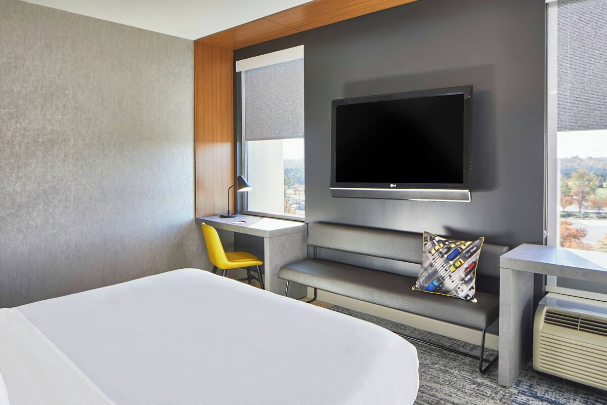 A modern hotel room with a large bed, a desk with a chair, a flat-screen TV mounted on the wall, and a window offering a city view.