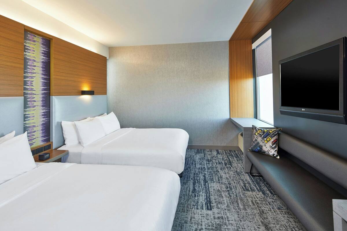 A hotel room with two beds, a large flat-screen TV, a wall-mounted sound system, and a window with a view.