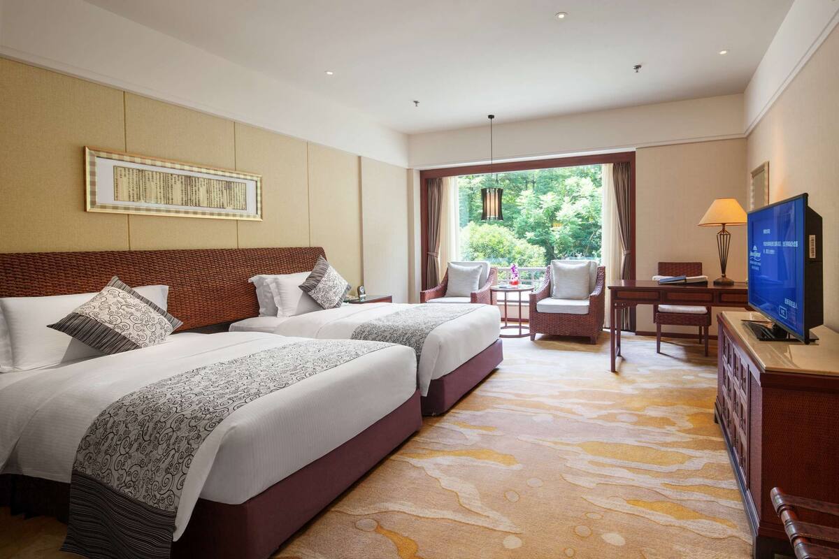 A hotel room with two beds, a television, a desk, and a view of greenery outside.