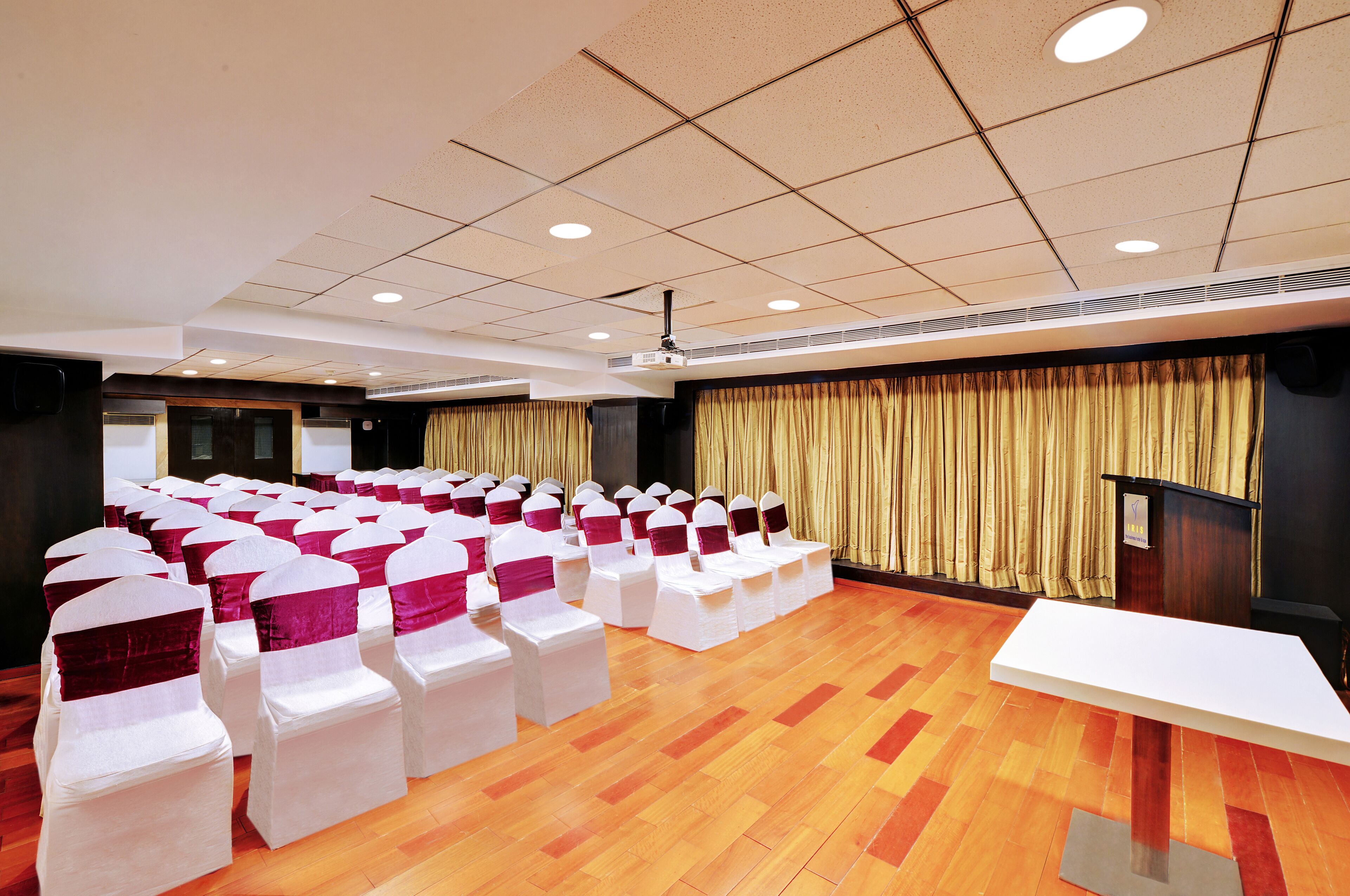 A conference room with rows of white chairs with red sashes, a podium, and a wooden floor.