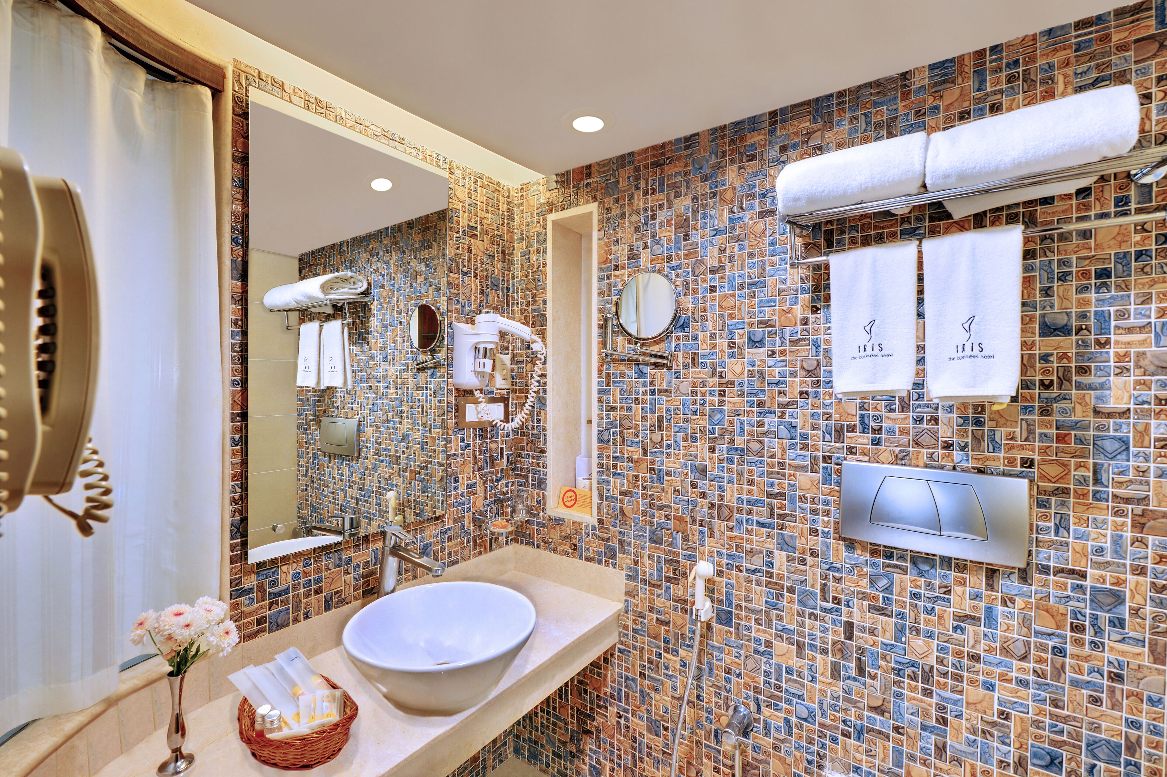 Un baño con pared de mosaicos, un espejo grande, un lavabo y toallas colgadas en un perchero.