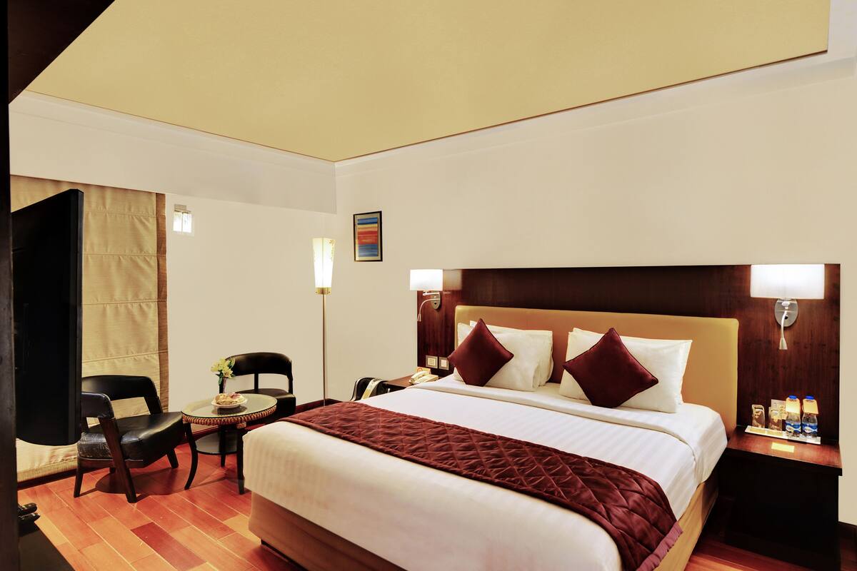 A hotel room with a large bed, a desk with a chair, a small table with a vase of flowers, and a nightstand with a lamp and a bottle.