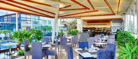 A spacious outdoor dining area with multiple tables and chairs, surrounded by large windows and potted plants.