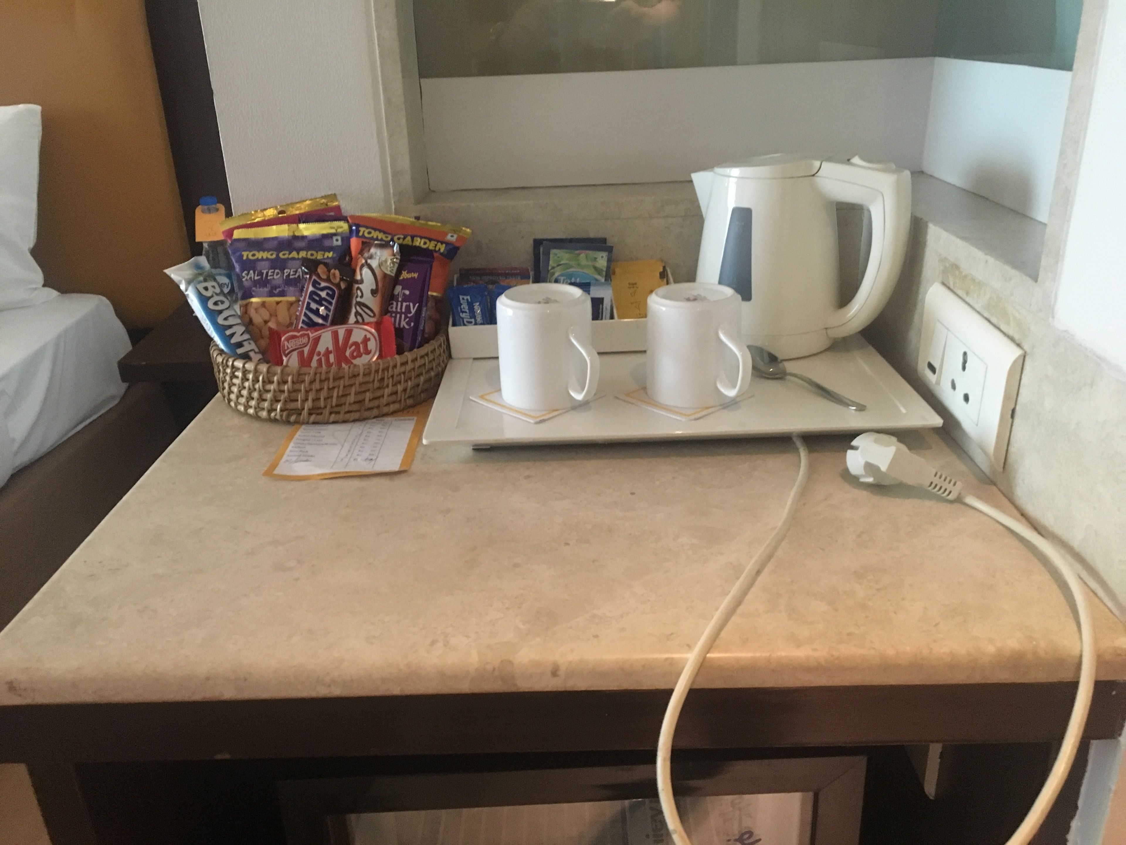 A hotel room with a kitchenette area. There is a basket of snacks, a kettle, and two white mugs on a tray.
