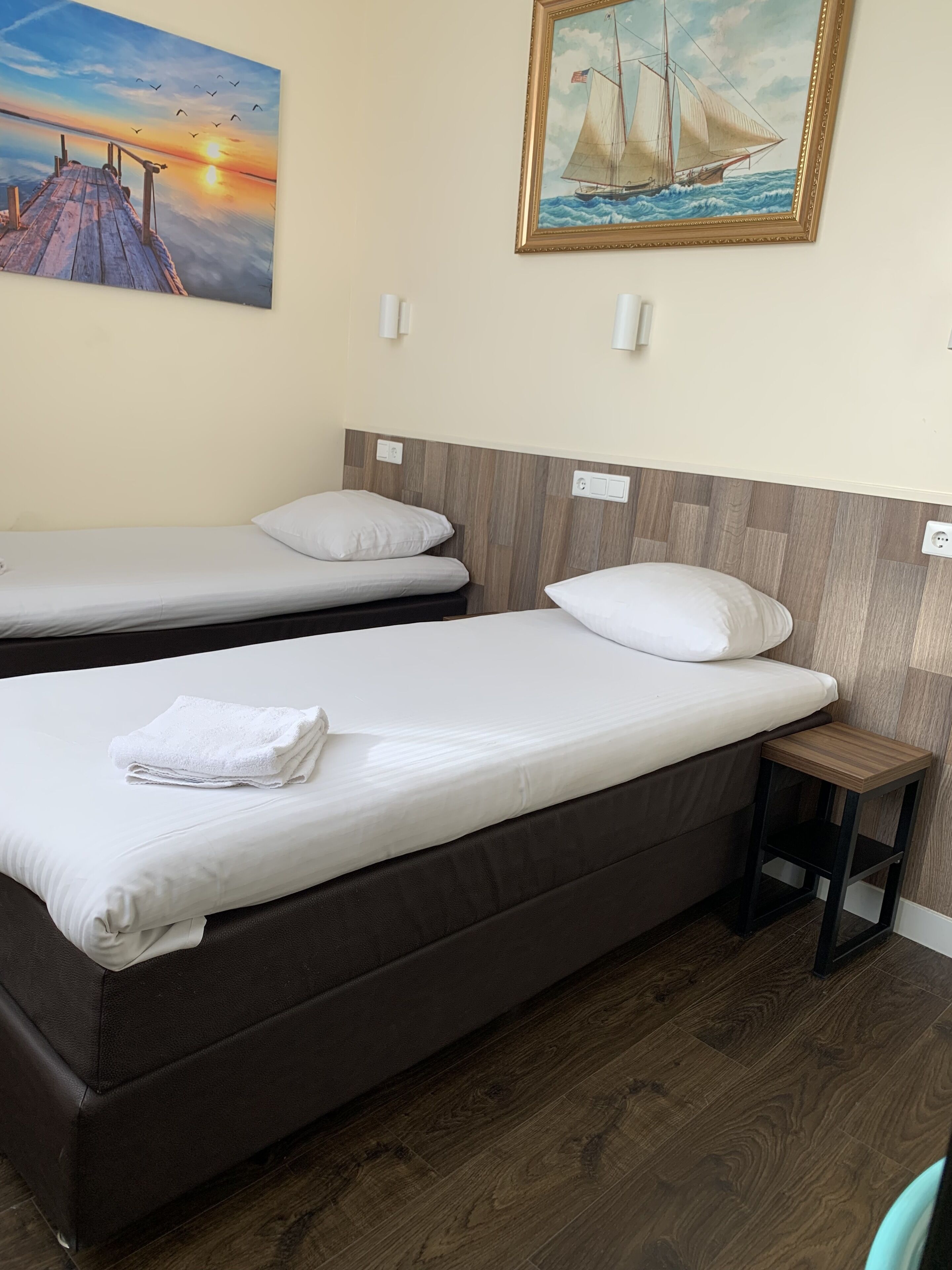 A hotel room with two beds, a wooden headboard, a small bedside table, and a painting of a sailboat on the wall.