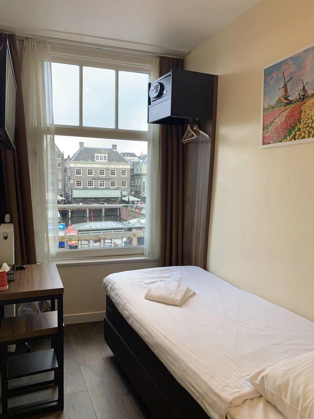 A hotel room with a bed, a desk, a TV mounted on the wall, and a window with a view of buildings and a street.