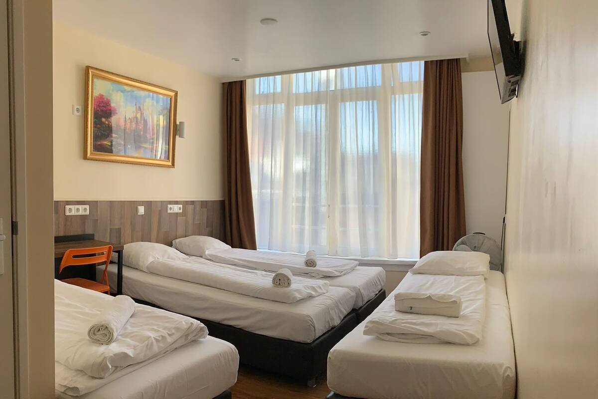 A hotel room with two beds, a desk, a chair, a painting on the wall, and a large window with curtains.