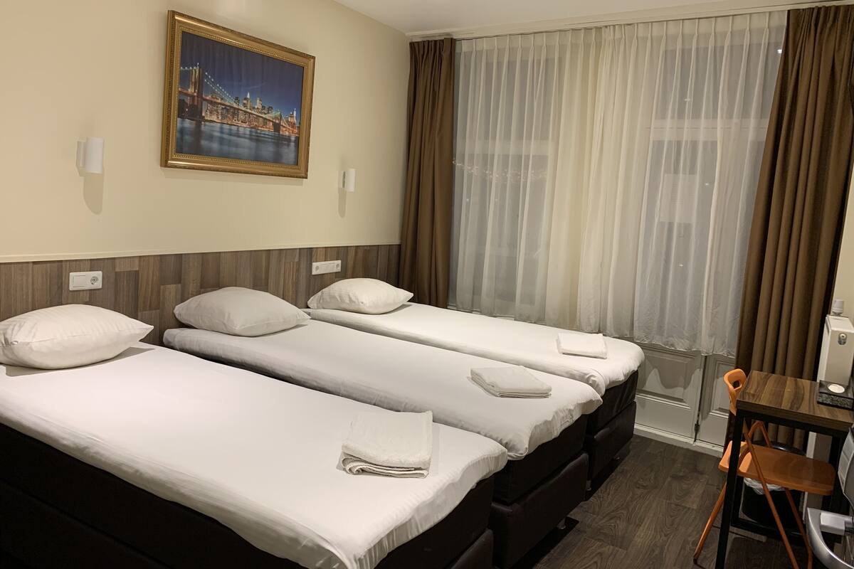 A hotel room with two beds, a wooden desk, a chair, a painting of a bridge, and sheer curtains.