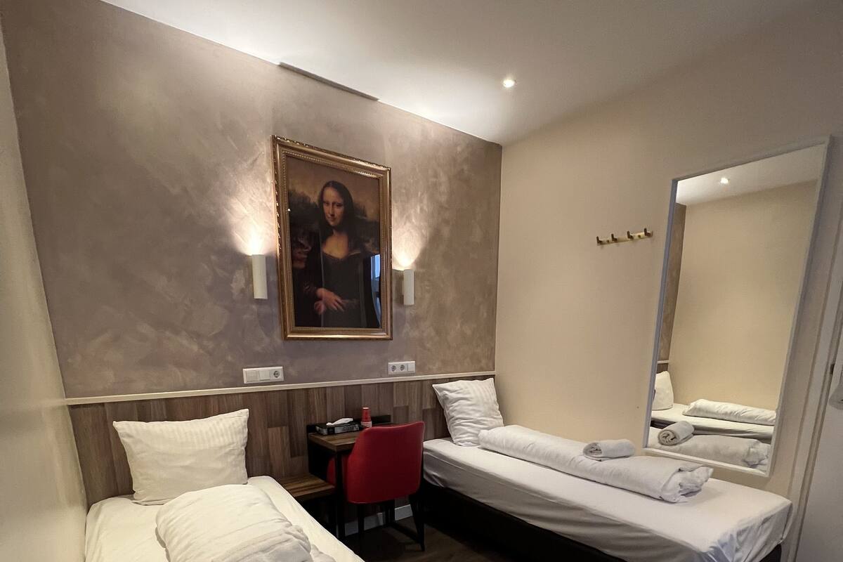 A hotel room with two beds, a desk, a chair, a painting on the wall, and a mirror.