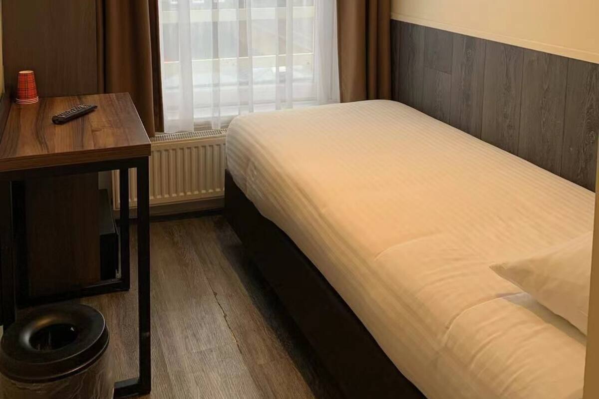 A hotel room with a bed, a small table with a remote control, a radiator, and a view of buildings through sheer curtains.