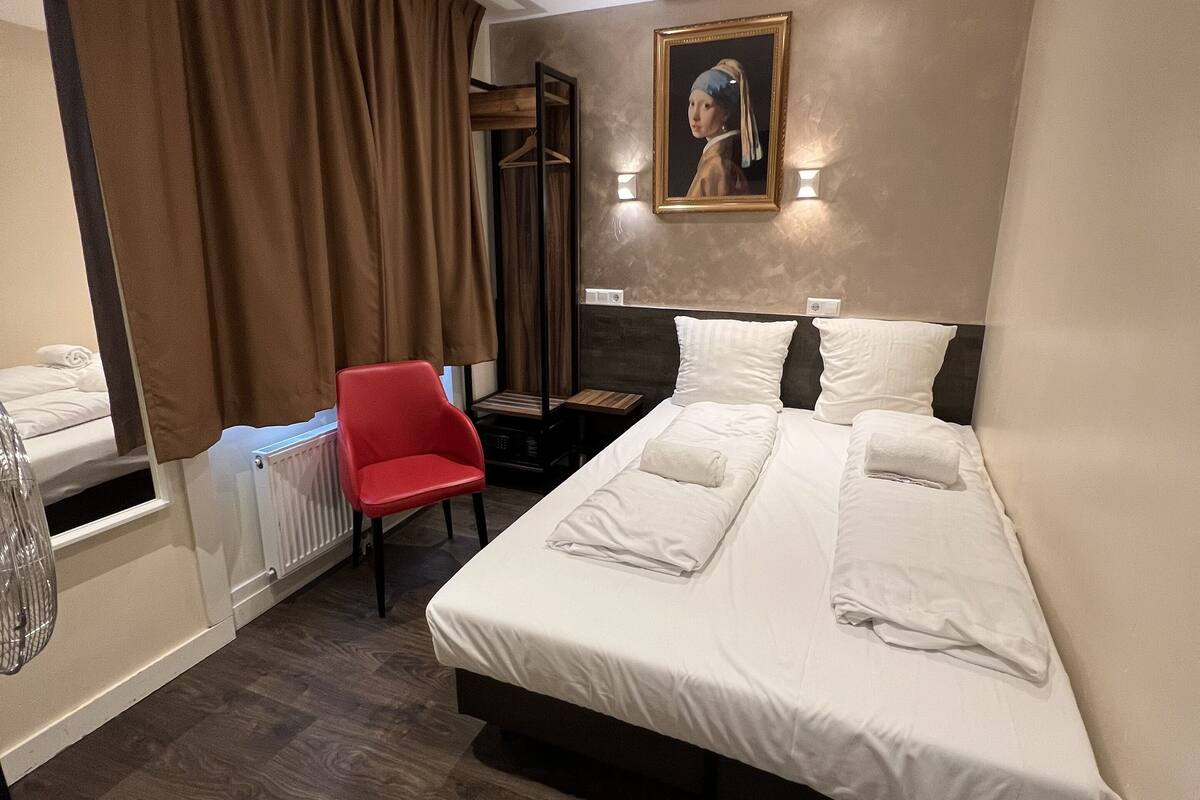 A hotel room with a bed, a red chair, a desk, and a painting on the wall.