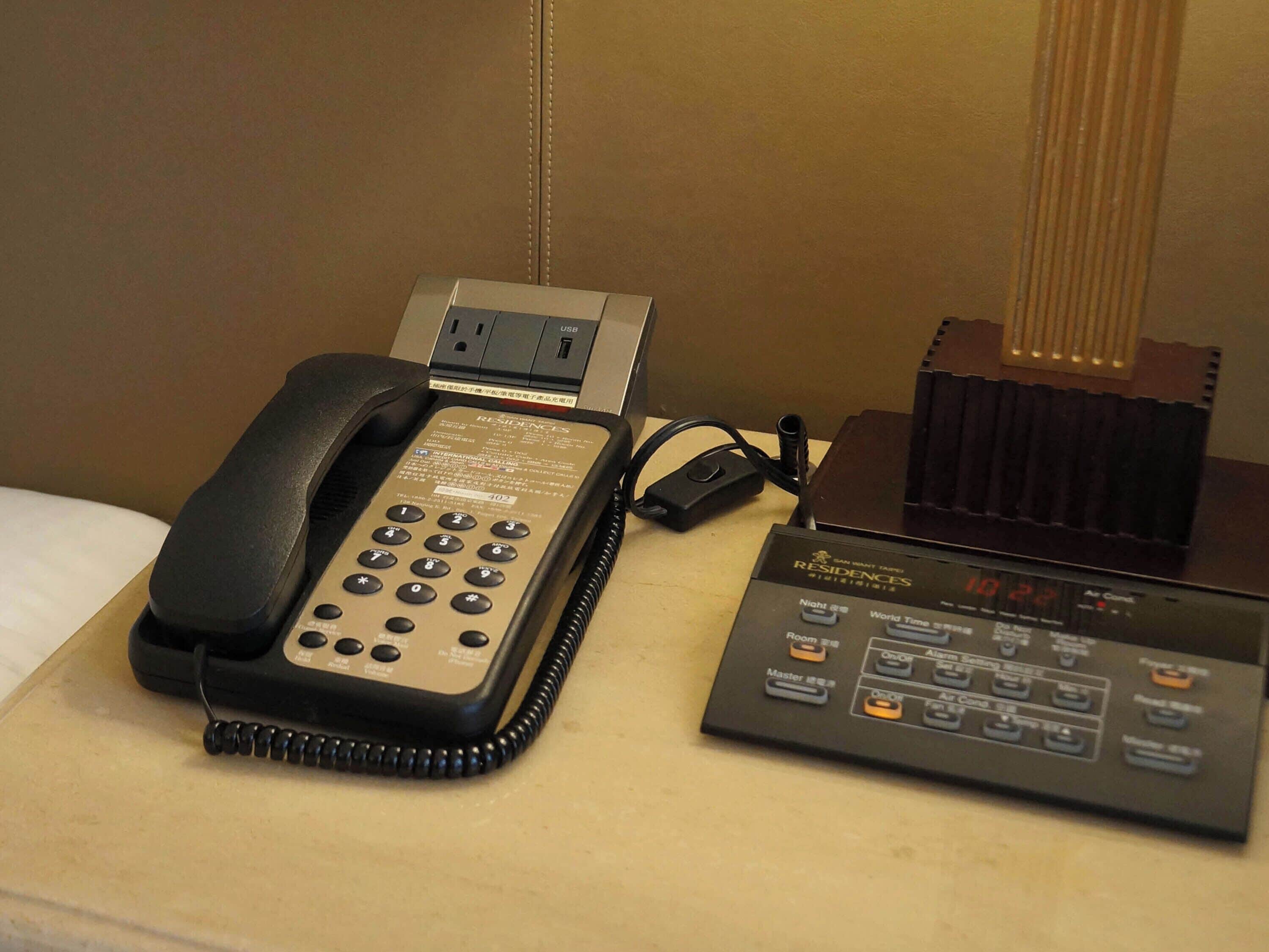 Quarto de hotel com telefone, régua de tomadas e painel de controle.