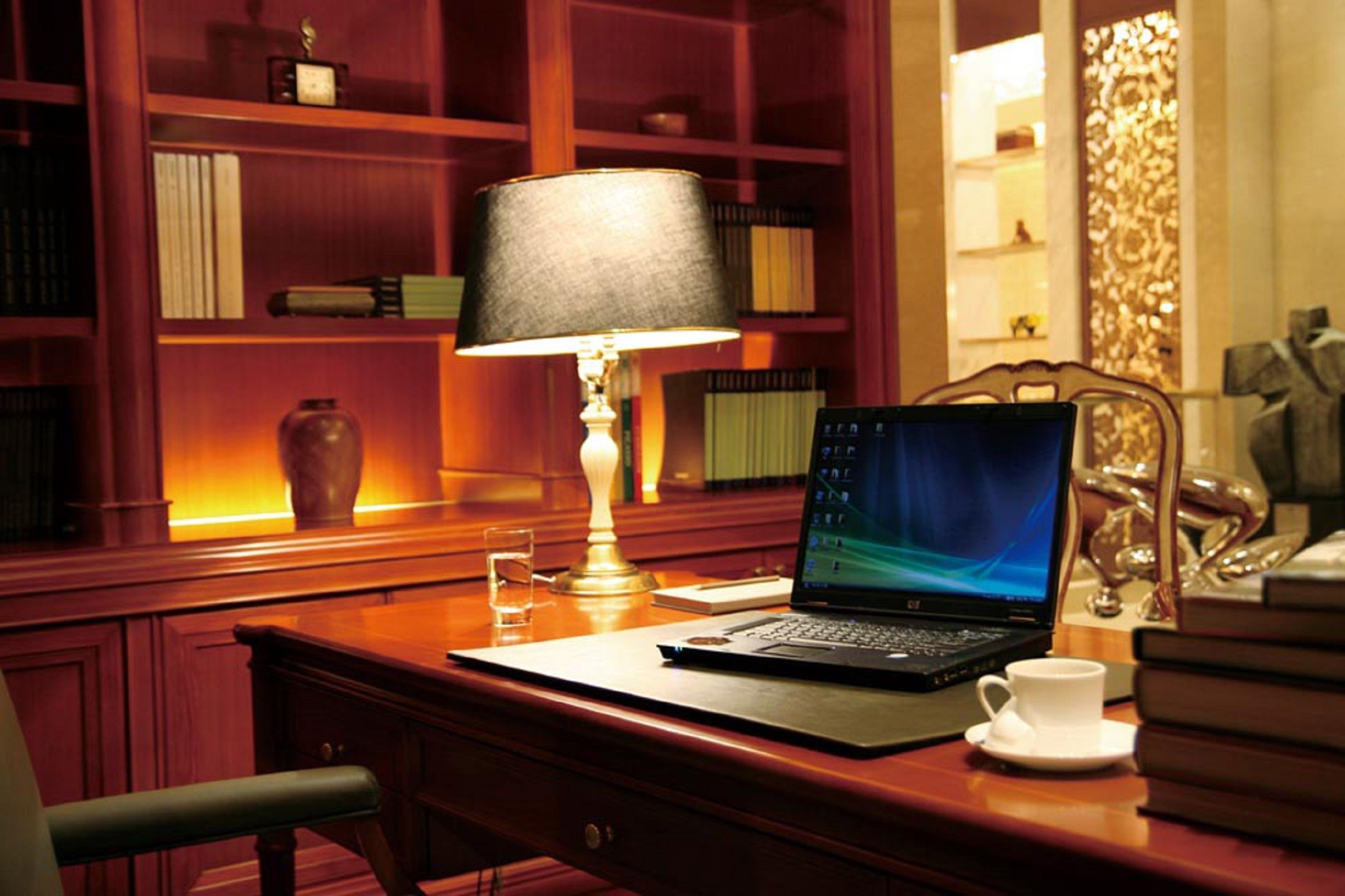 A wooden desk with a laptop, a lamp, a cup, and a bookshelf in the background.