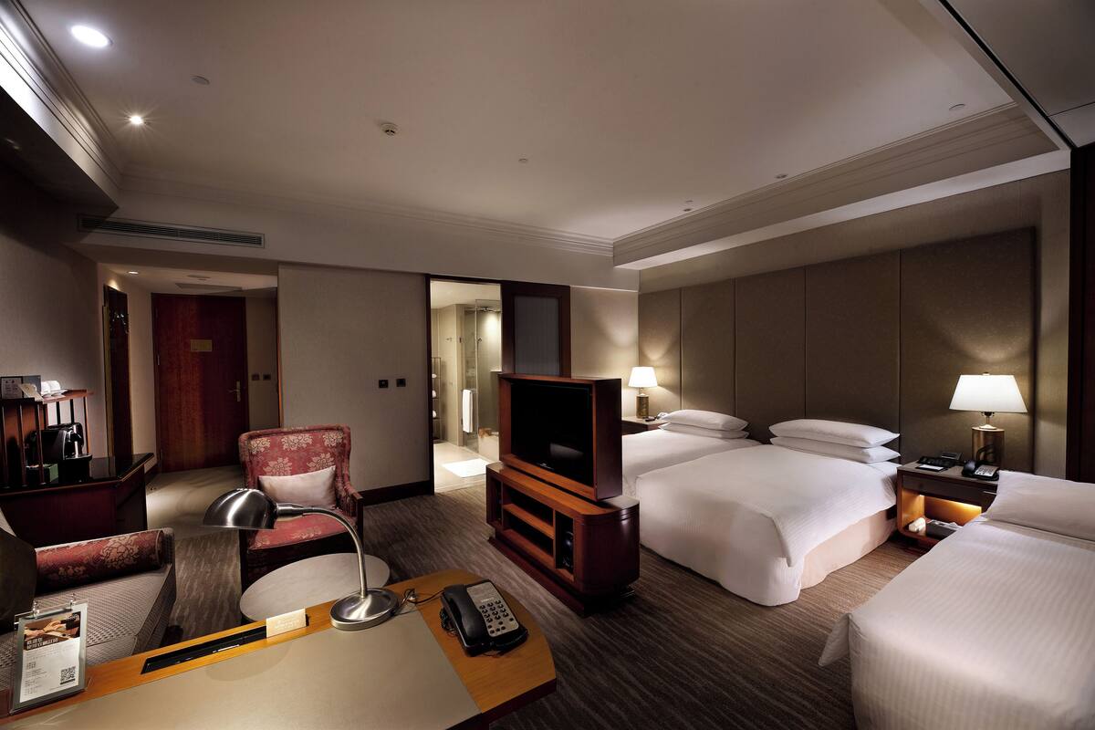 A hotel room with a bed, a TV, a desk with a lamp, a chair, and a bedside table with a lamp.