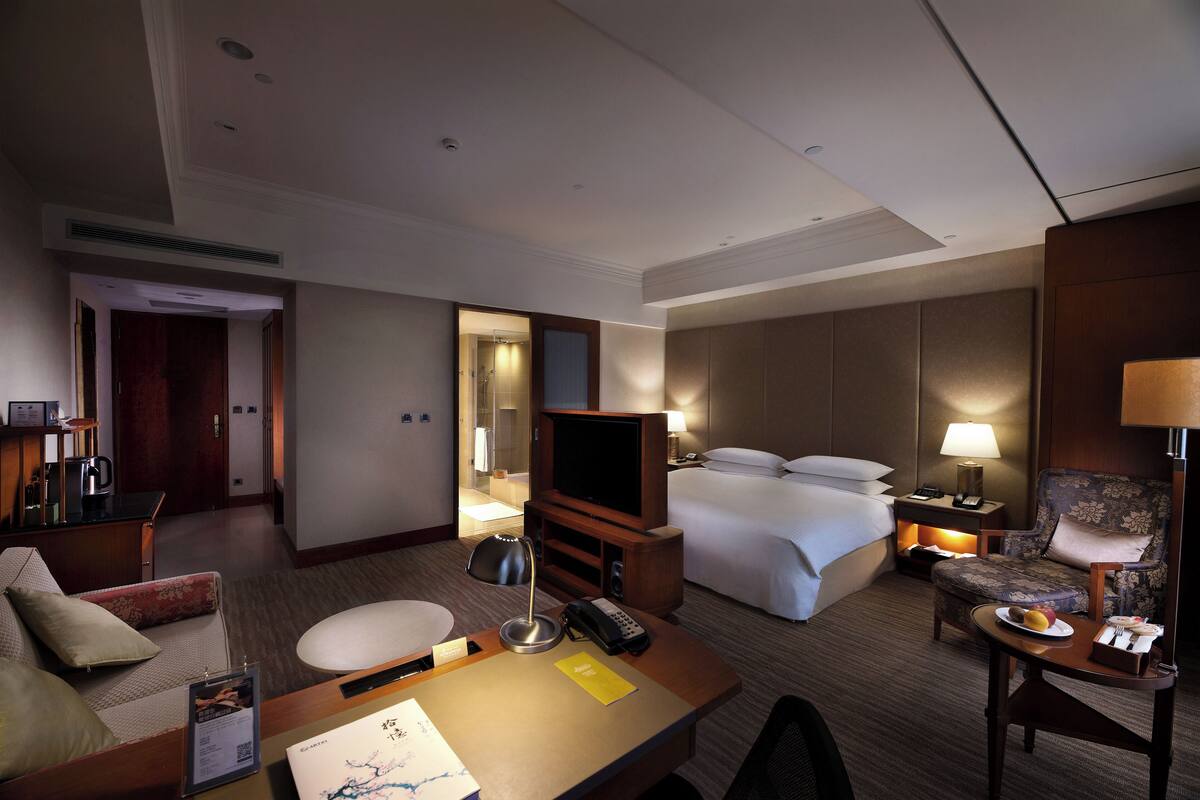 A hotel room with a bed, a desk with a lamp, a television, a chair, and a small table with a plate of fruit.