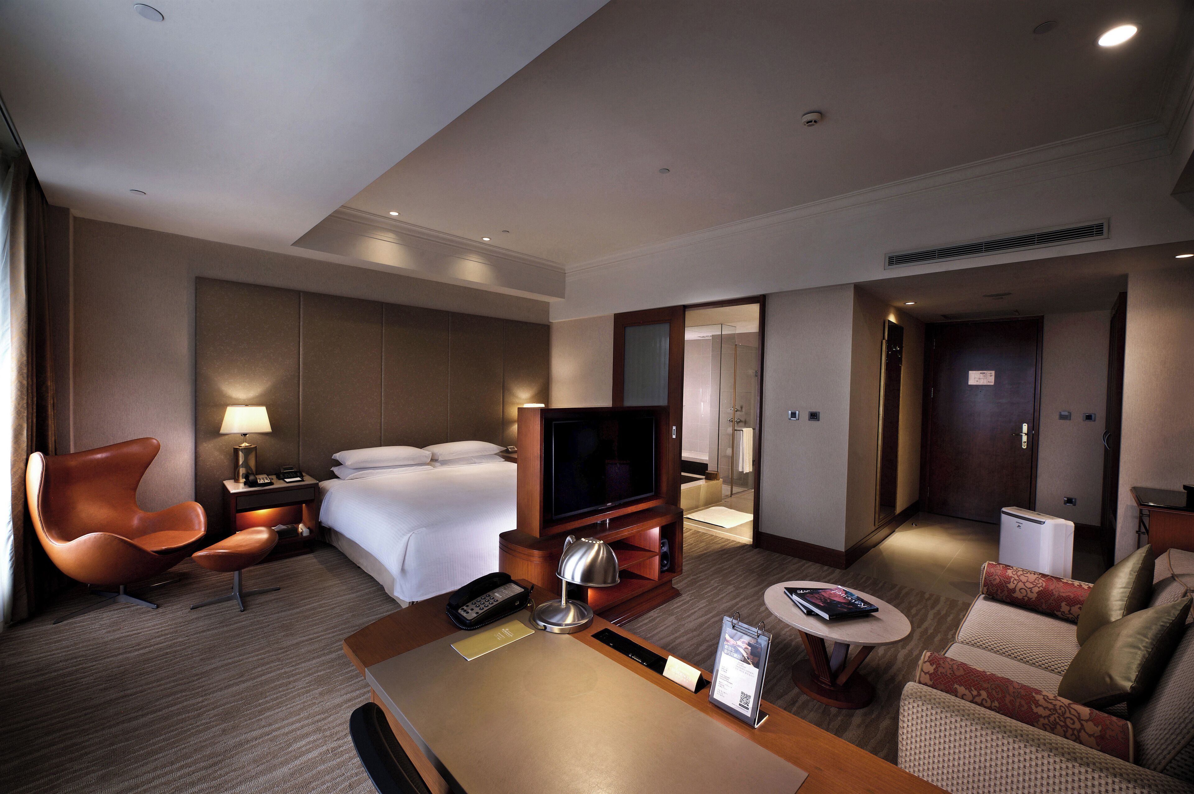 A hotel room with a bed, a TV, a desk with a lamp, a chair, and a sofa.