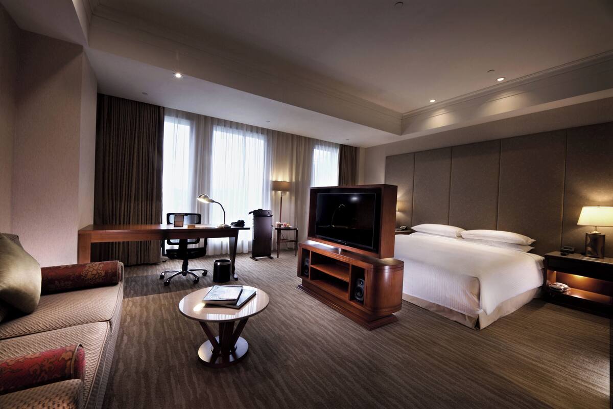 A hotel room with a bed, a TV, a desk, a chair, a coffee table, and a lamp.