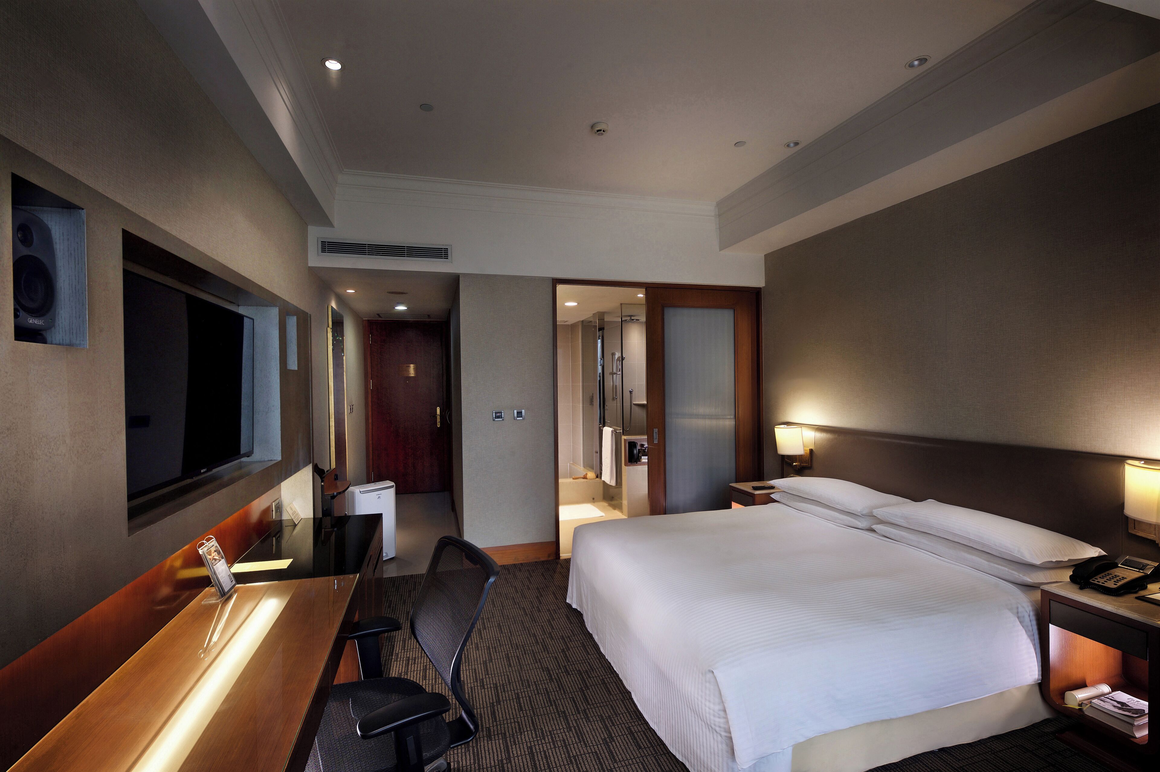 Une chambre d’hôtel avec un grand lit, un bureau, une chaise, une télévision, un miroir et une salle de bain visible à travers une porte ouverte.