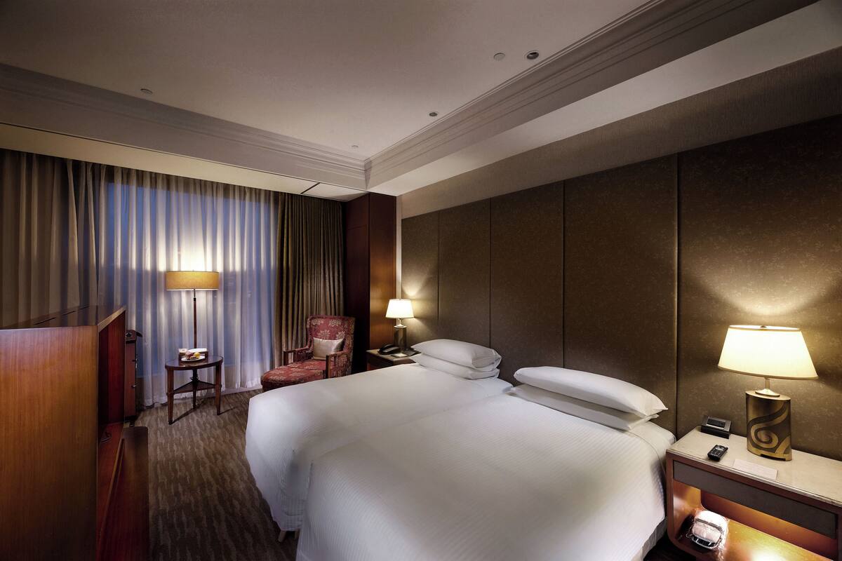 A hotel room with a large bed, a desk with a lamp, a chair, and a wardrobe.
