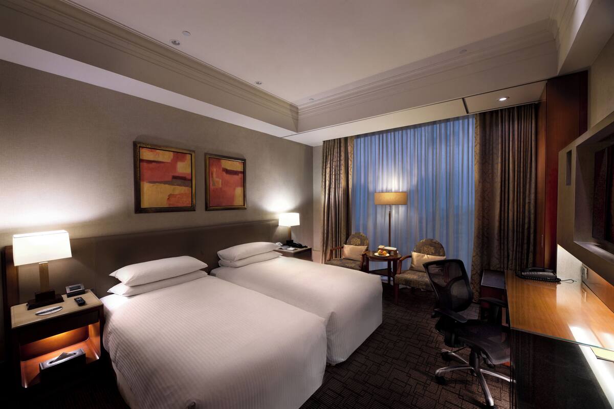 A hotel room with two beds, a desk, a chair, a TV, and a window with curtains.