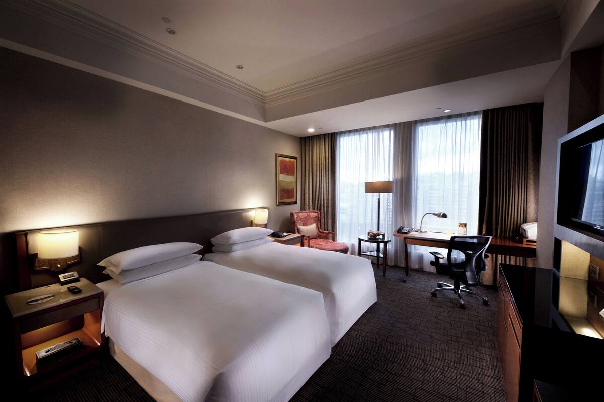 A hotel room with two beds, a desk, a chair, a TV, and a window with curtains.