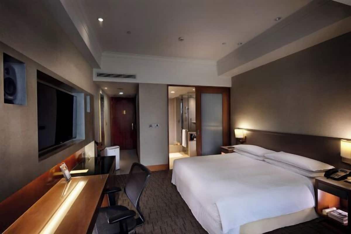 A hotel room with a large bed, a desk with a chair, a TV, and a bathroom visible through an open door.