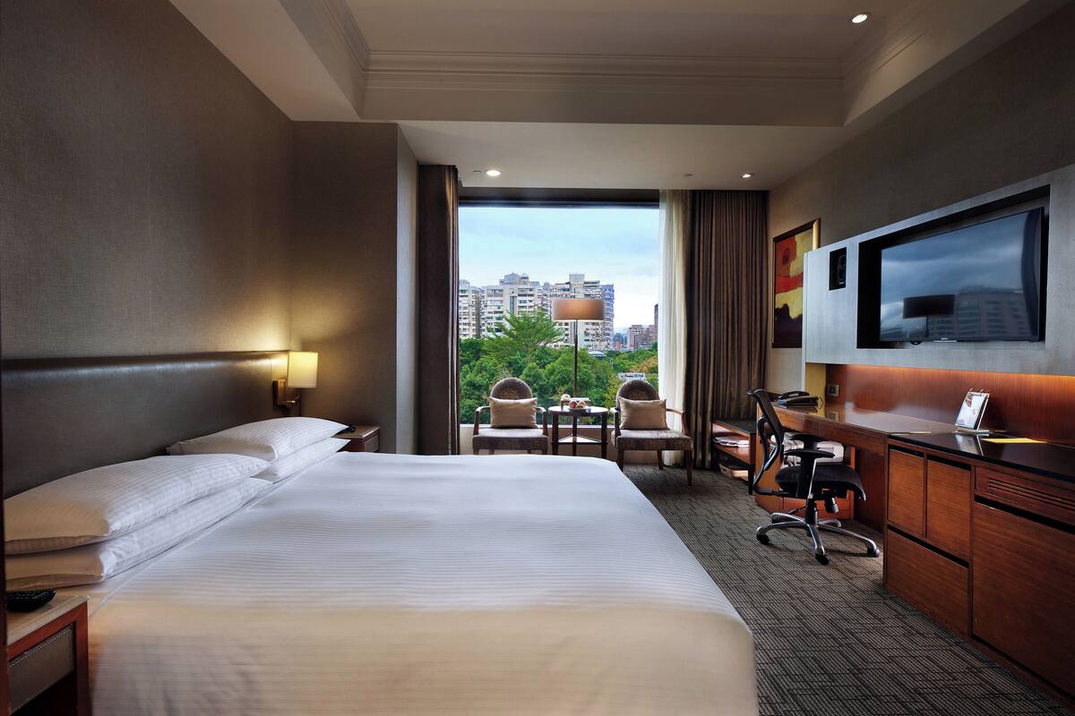 A hotel room with a large bed, a desk with a chair, a TV, and a view of buildings through the window.