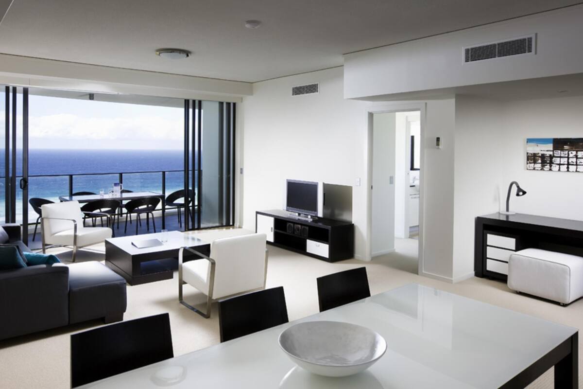 Ein modernes Wohnzimmer mit Balkon und Meerblick, einem Essbereich mit weißem Tisch sowie einer Küche mit schwarzer Arbeitsplatte.