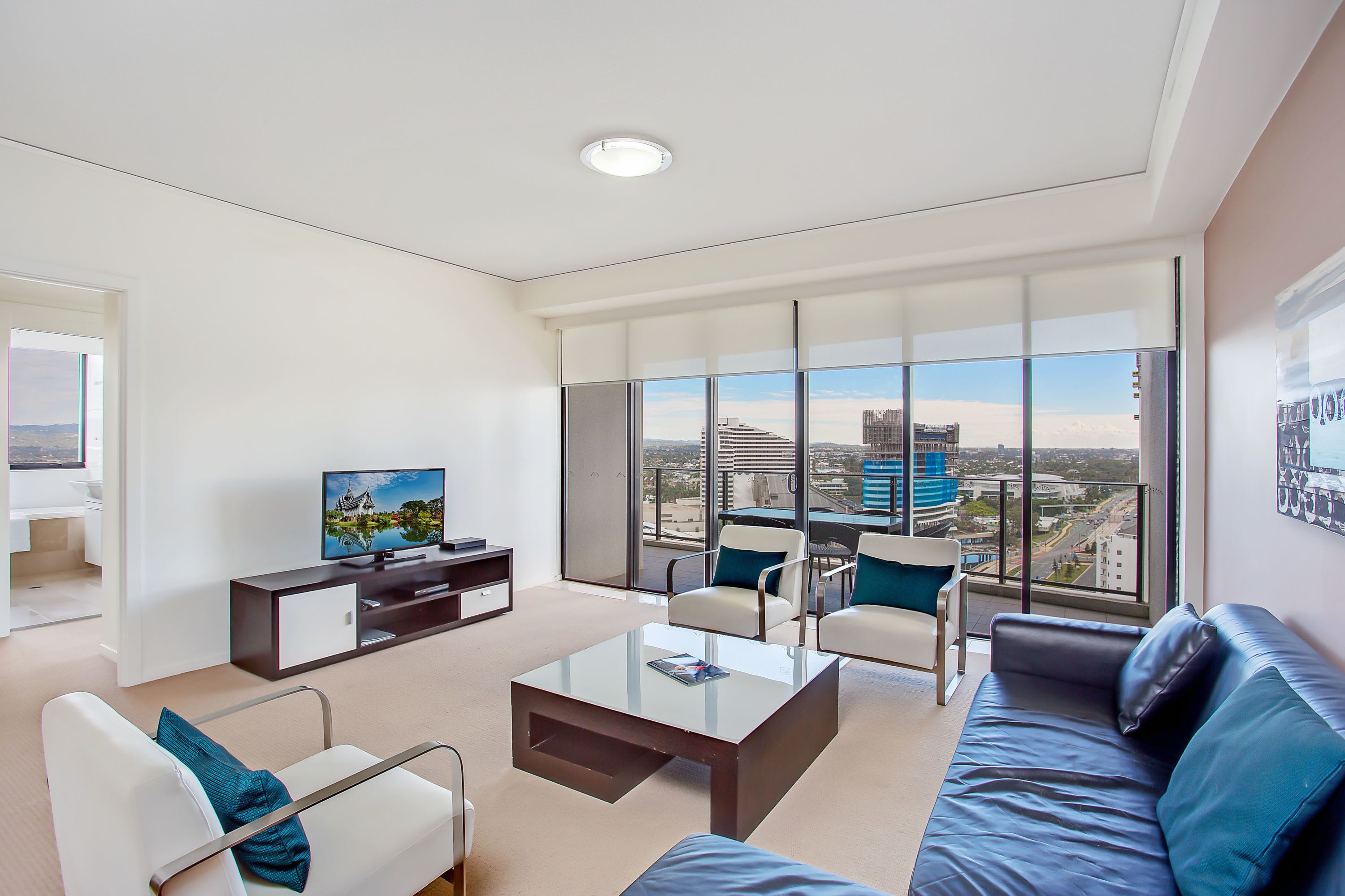 Ein modernes Wohnzimmer mit Flachbildfernseher, Glastisch, Sofa und Sessel. Große Fenster bieten Blick auf die Stadt.