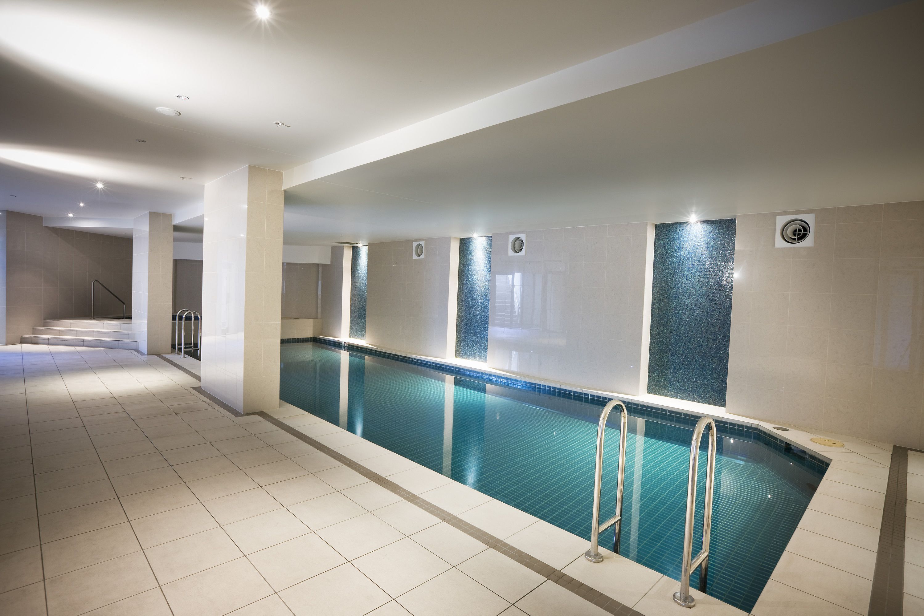 A swimming pool with tiled walls and a tiled floor, surrounded by a corridor with a staircase leading to an upper level.