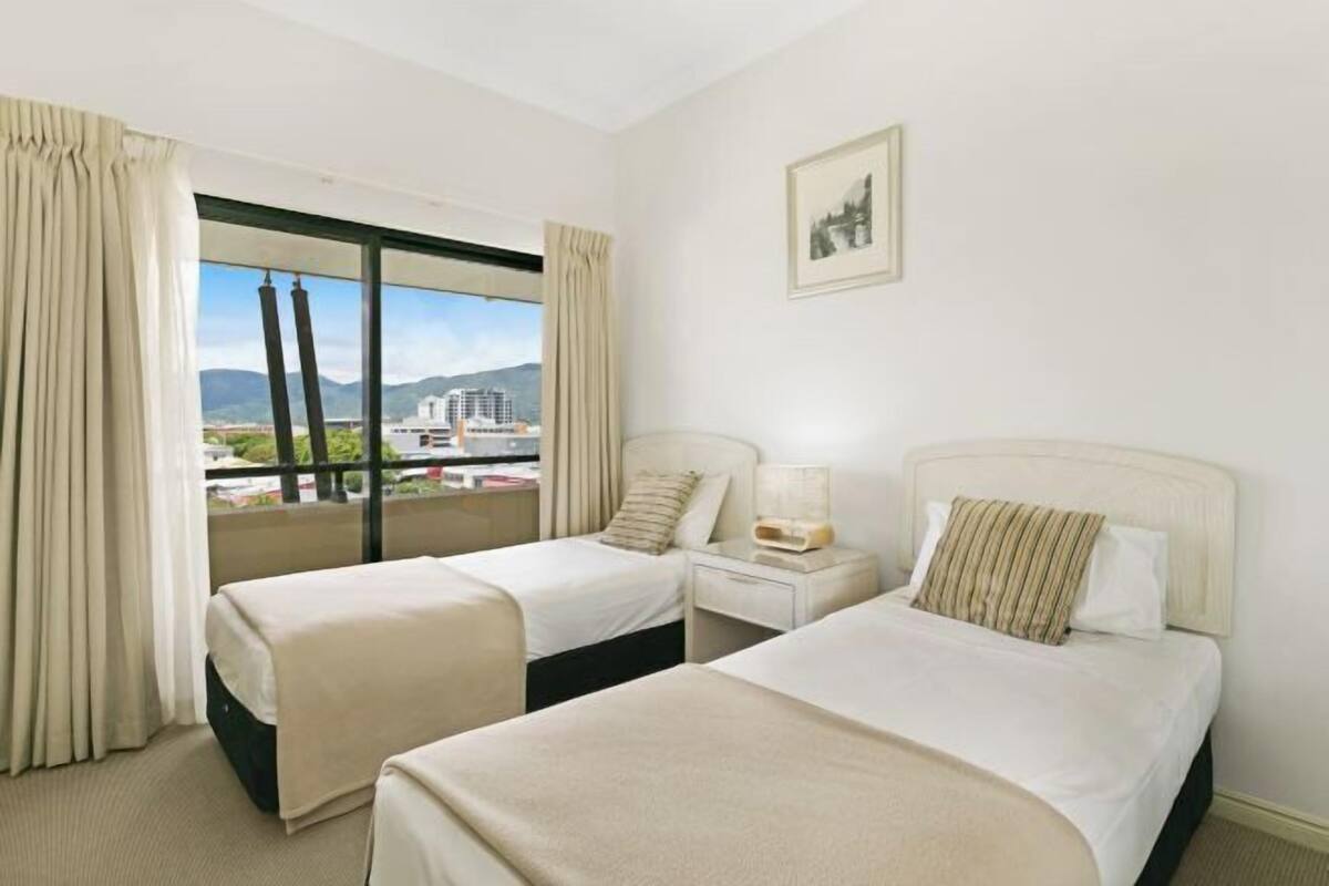 Ein Hotelzimmer mit zwei Betten, einem Fenster mit Vorhängen, einem Nachttisch, einer Lampe und einem gerahmten Bild an der Wand.