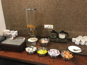 Disposição de buffet de café da manhã com diversos alimentos, pratos descartáveis e copos.