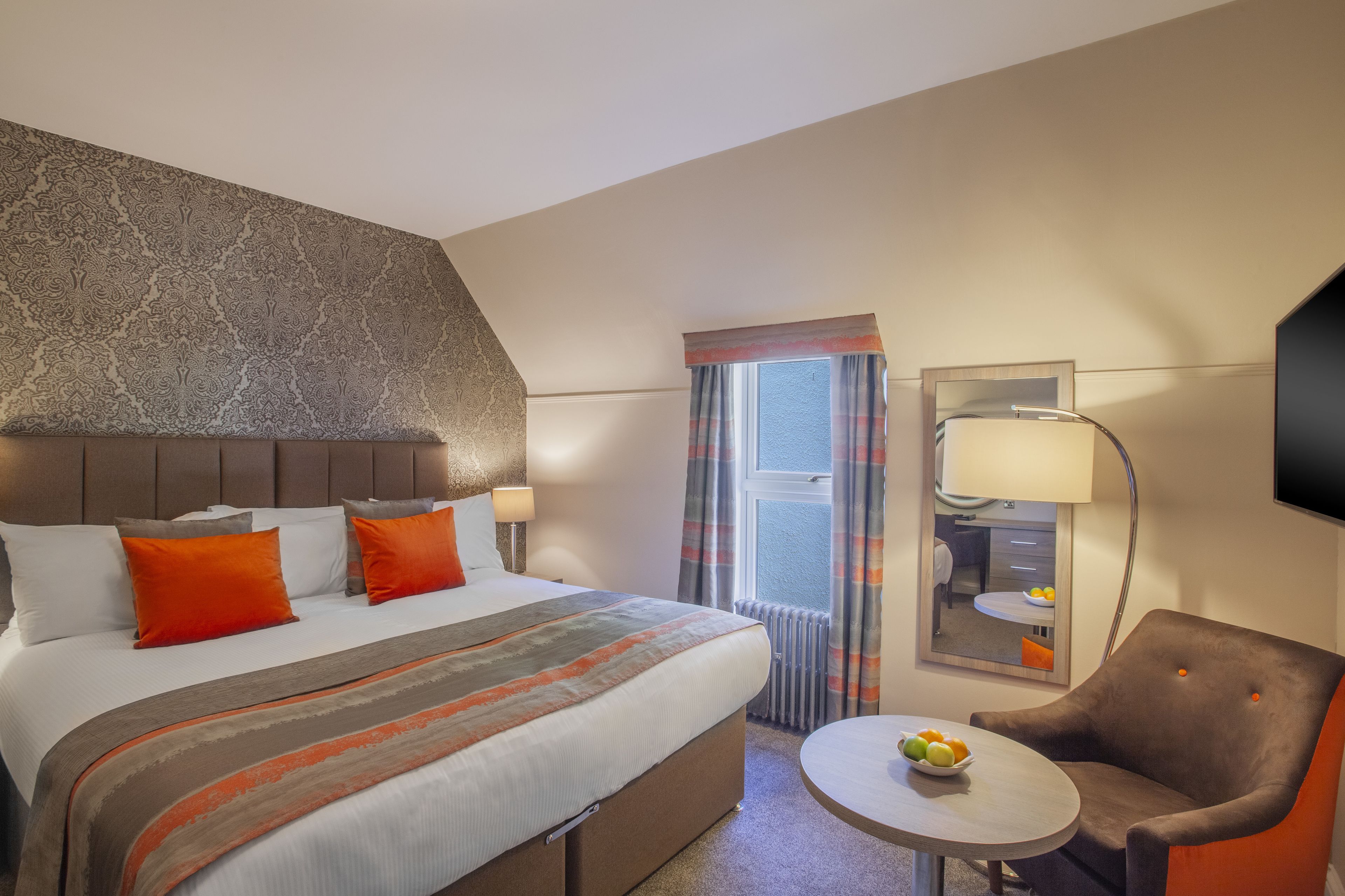 Una camera d'albergo con un letto grande, un tavolo rotondo con frutta, una sedia, uno specchio e una televisione a parete.