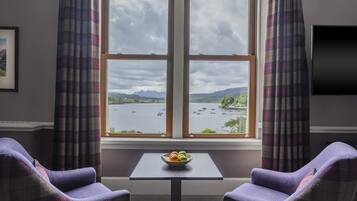 A room with a large window, two purple armchairs, a small table with a bowl of fruit, and a flat-screen TV on the wall.
