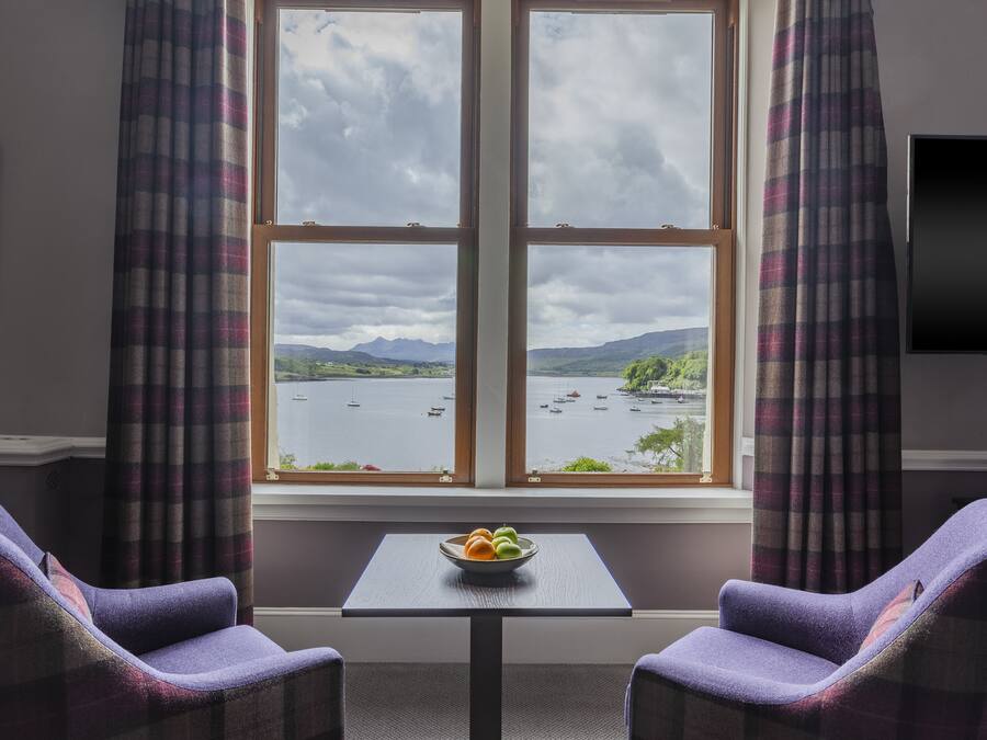 A room with a large window, two purple armchairs, a small table with a bowl of fruit, and a flat-screen TV on the wall.