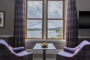 A room with a large window, two purple armchairs, a small table with a bowl of fruit, and a flat-screen TV on the wall.