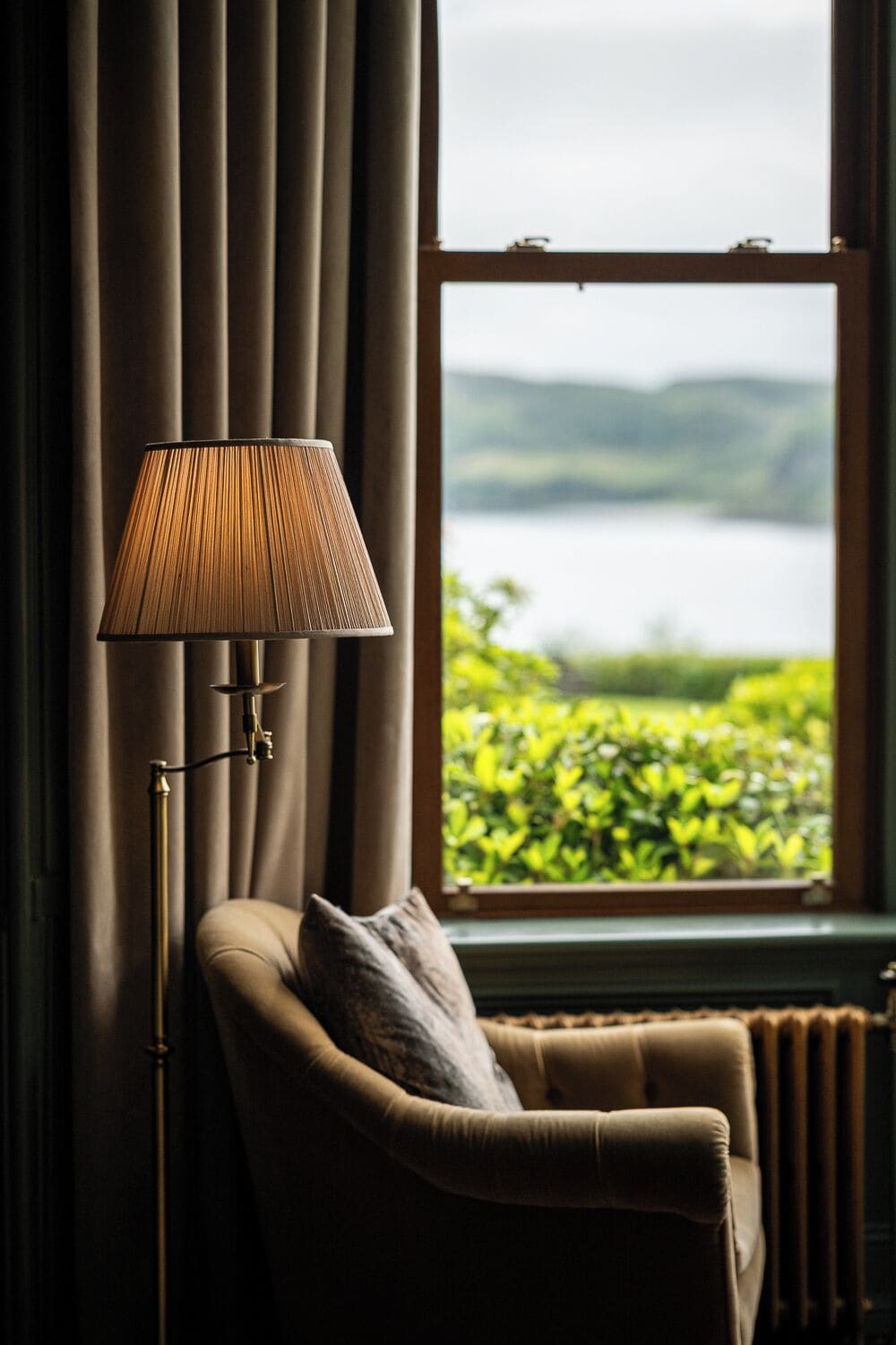 A room with a lamp, a chair, and a view of a lake through a window.
