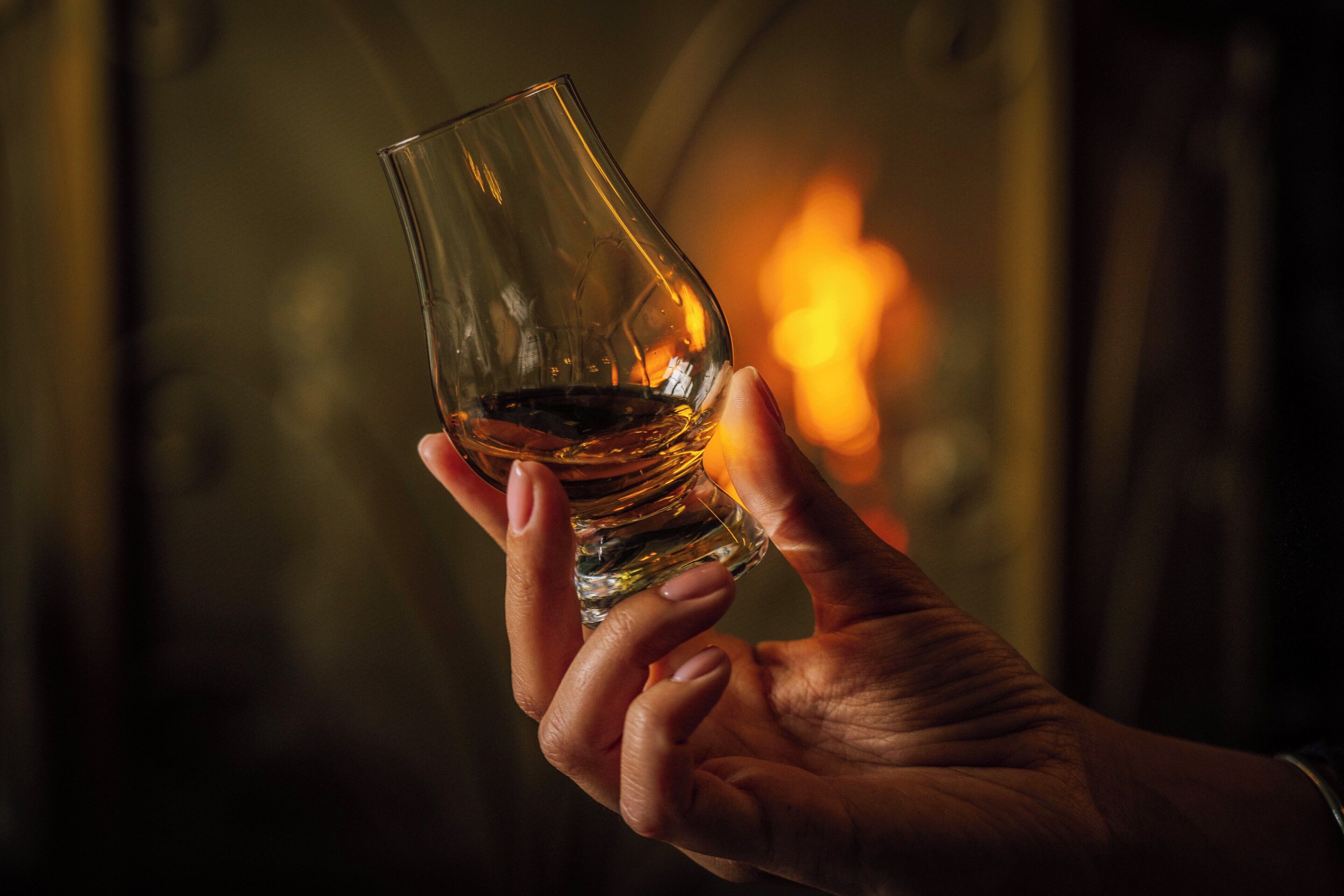 A hand holding a glass with a liquid, set against a blurred background with a warm light source.