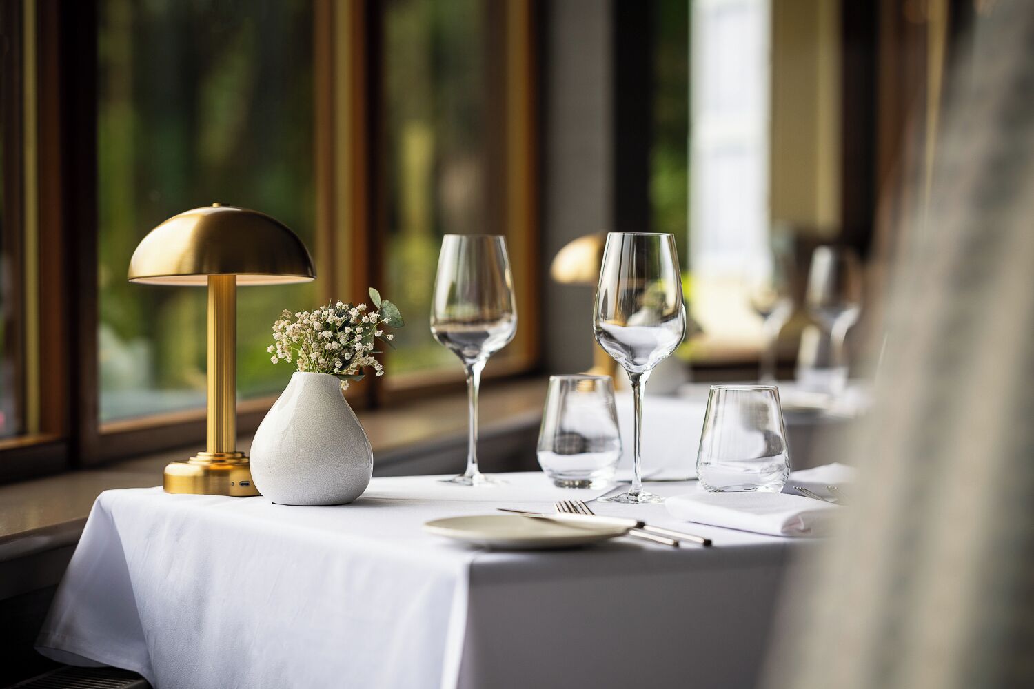 Une table à manger dressée avec une nappe blanche, une lampe dorée, un vase rempli de fleurs et des verres à vin.