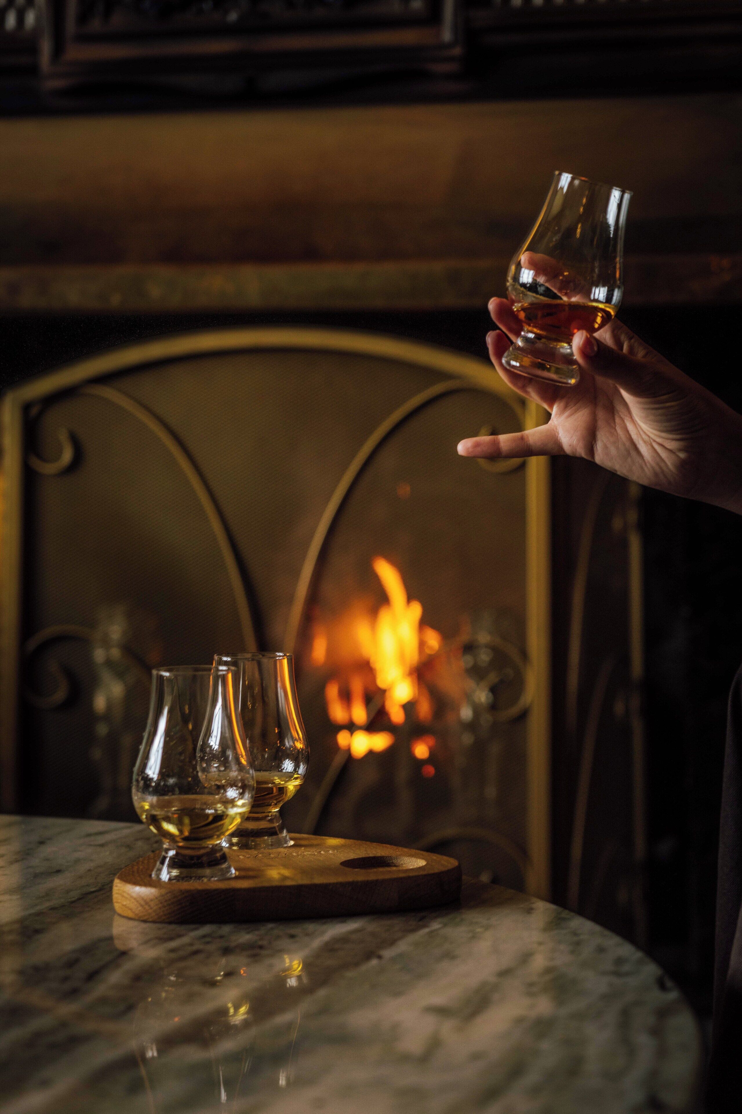 Une main tenant un verre de whisky, avec deux autres verres posés sur un dessous de verre, devant une cheminée.