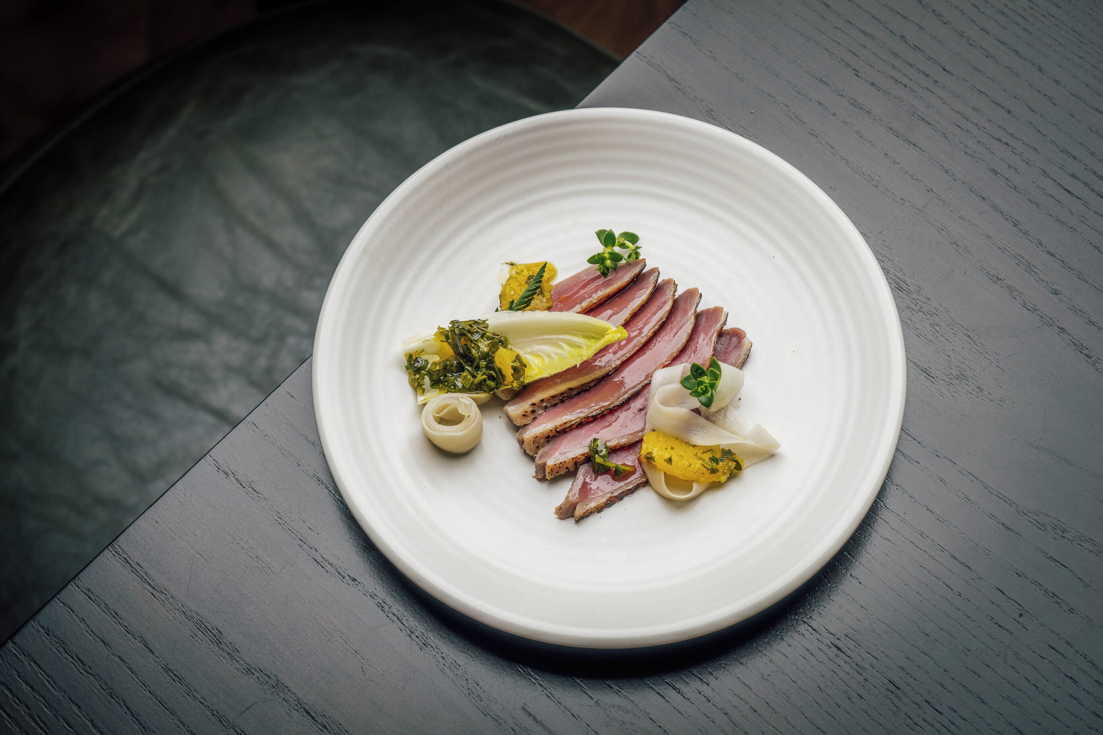 A white plate with sliced meat, vegetables, and a small white object on a dark wooden table.