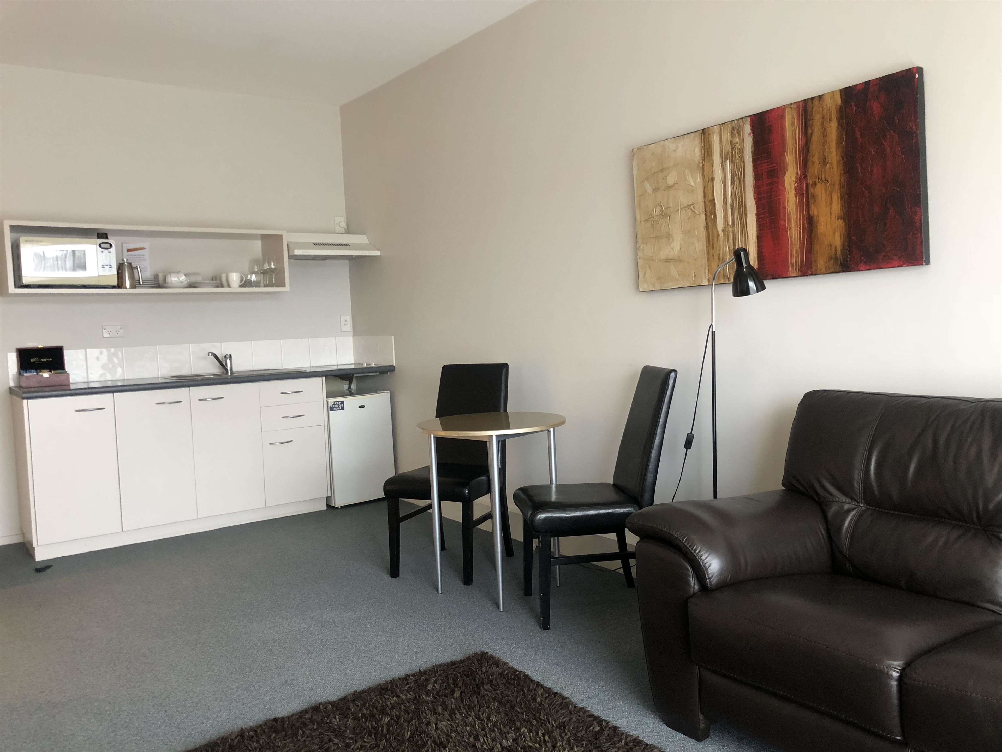 A modern living room with a brown leather sofa, a small round table, two black chairs, a kitchen area with white cabinets and a microwave, and a large abstract painting on the wall.