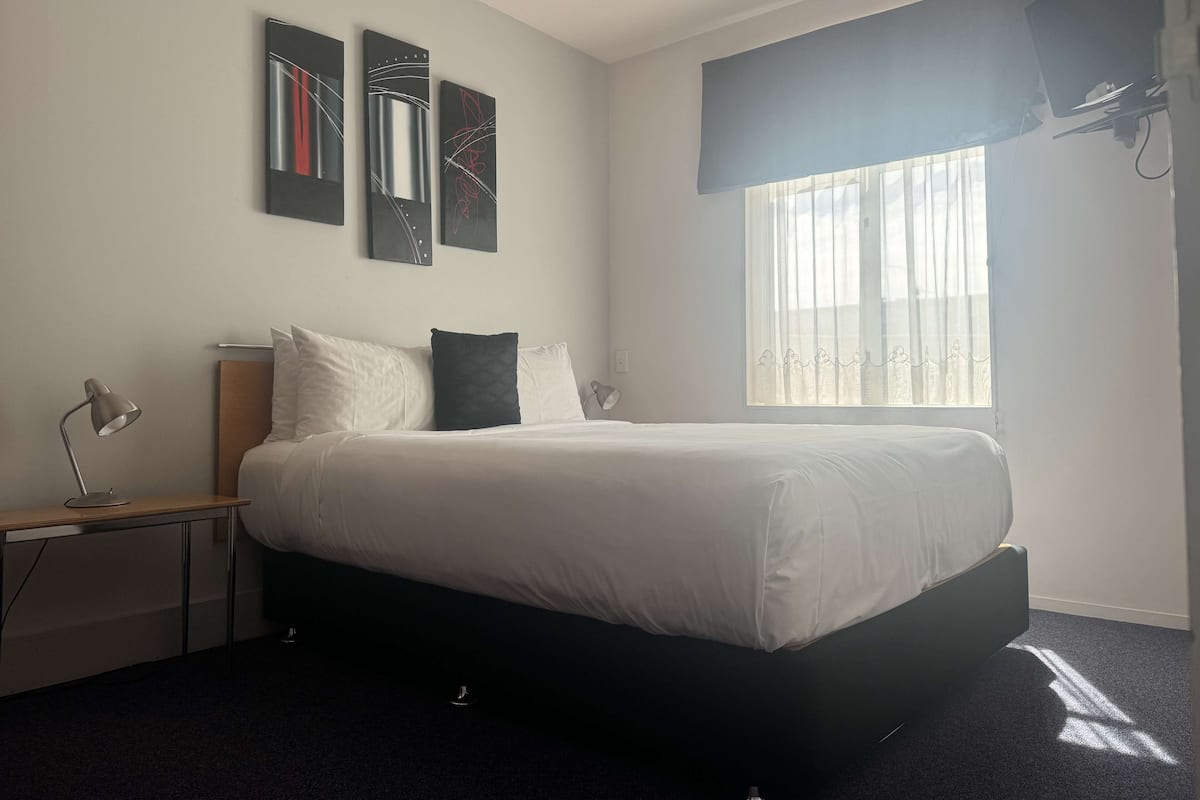 A modern hotel room with a large bed, a desk with a lamp, and artwork on the wall.