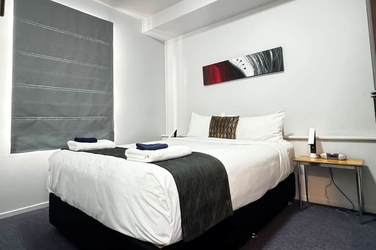 A modern hotel room with a large bed, a bedside table with a lamp, and a window with blinds.